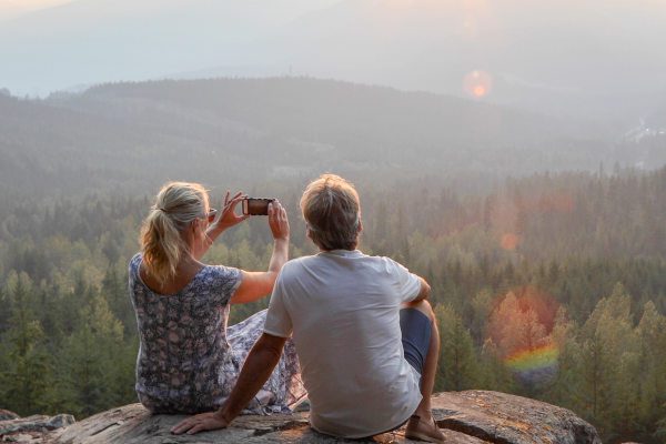 Retired couple sitting on mountain ledge taking photo of valley of trees and mountain range below them