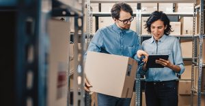 Female Inventory Manager Shows Digital Tablet Information to a Worker Holding Cardboard Box, They Talk and Do Work. In the Background Stock of Parcels with Products Ready for Shipment.