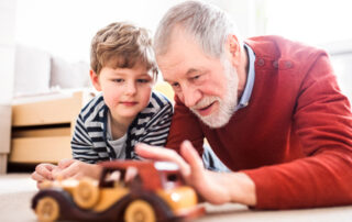 An elderly man | On his right is his grandson | They are both laying on the floor | The are playing with a toy car