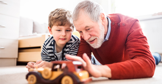 An elderly man | On his right is his grandson | They are both laying on the floor | The are playing with a toy car
