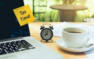 A coffee table | On top of it to the right are three pieces of paper | towards the center on top of the papers is a plate with a cup of coffee on it | to the right of the coffee is a small alarm clock | the clock is black and reads 8:03 am | to the right of the clock is a MacBook | the MacBook is turned off with a yellow sticky note of it that says Tax Time! |