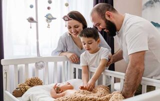 In a well lit room | A family of three | A mother, child, and father| They look over a crib | There a new born rests |