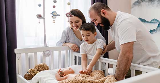 In a well lit room | A family of three | A mother, child, and father| They look over a crib | There a new born rests |